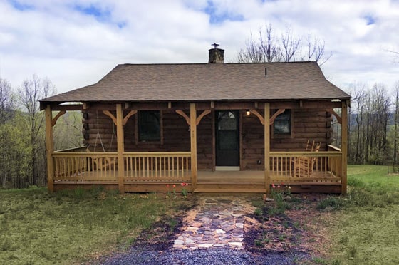 A rustic cabin with log construction is presented, featuring a spacious front porch with rocking chairs. A stone pathway leads to the entrance, surrounded by lush greenery. The roof is sloped, and a chimney is visible at the center, complementing the cabin's natural setting.