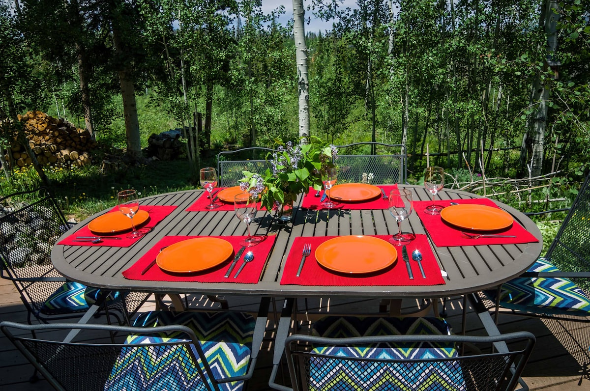 An outdoor dining set is positioned on a wooden deck, surrounded by lush greenery. The table is arranged with orange plates and glassware atop red placemats. Colorful patterned cushions add comfort to the metal chairs, creating a welcoming space for meals in nature.