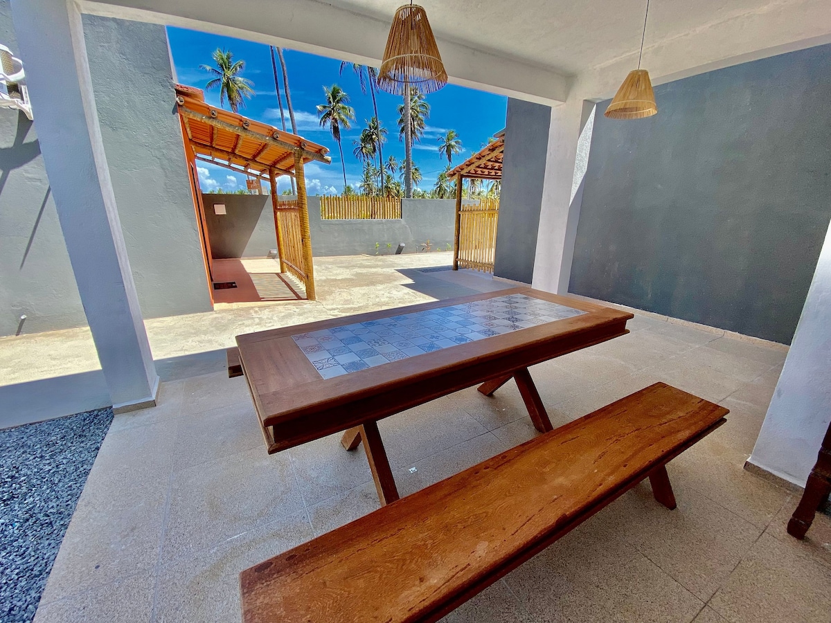 An outdoor dining area is presented with a wooden table and benches, designed for communal gatherings. The space is framed by palm trees and features natural light shining from a clear blue sky. Unique pendant lights hang from the ceiling, adding visual interest.