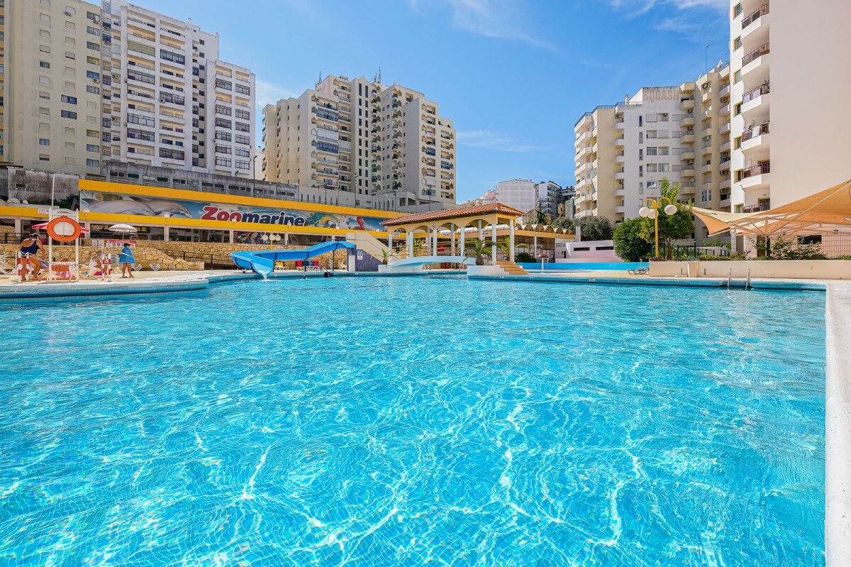 A large outdoor swimming pool is visible, surrounded by modern residential buildings. The clear water reflects the blue sky, and lounge areas can be seen along the pool's edge, inviting relaxation. Shade structures provide comfort for guests near the pool.