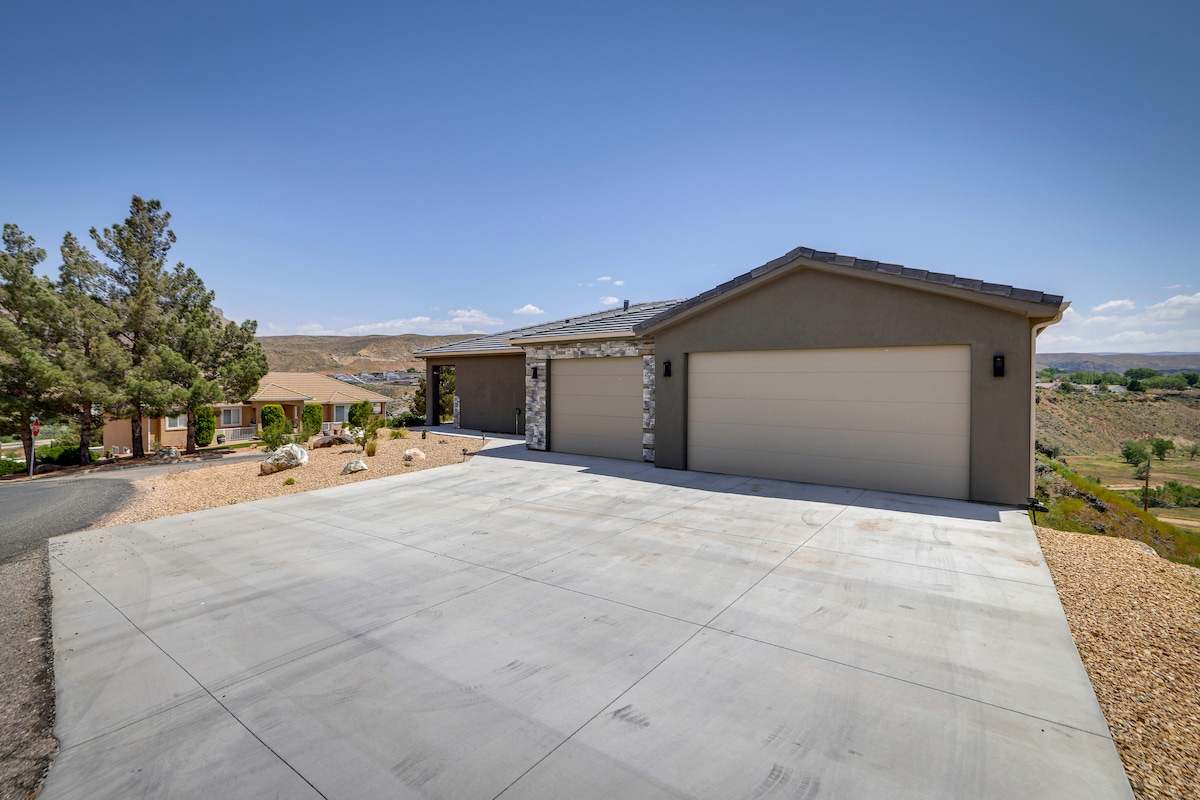 The exterior of the home showcases a modern design with a spacious driveway and attached garage. Surrounding landscaping includes low-maintenance rock features and mature trees. A scenic view of the surrounding hills and landscape is visible in the background under a clear blue sky.