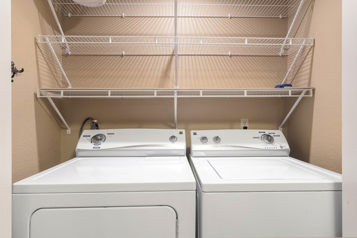 A laundry area showcases a pair of white appliances, a washer and dryer, positioned side by side. Above, a wire shelf provides space for storage. The wall features a warm, neutral color that complements the appliances.