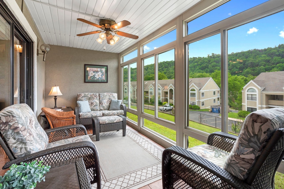 The all-season balcony features large windows that bring in natural light and offer views of the surrounding greenery. Comfortable seating options are arranged around a patterned area rug, with a small table and a decorative lamp adding a cozy touch to the space.