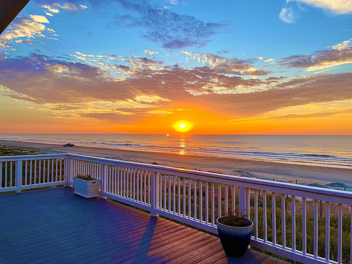 A vibrant sunset is captured over the ocean horizon, casting warm hues of orange and yellow across the sky. Gentle waves ripple along the shoreline, while a spacious deck with white railings provides a scenic vantage point for the tranquil beach view.