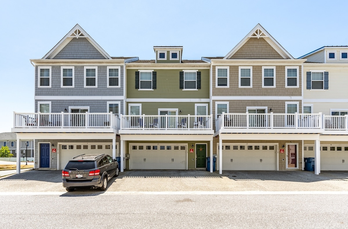 A row of modern townhouses features a mix of neutral-colored siding, including grey and green tones. Each townhouse has a covered front porch and open balcony, with garage doors visible below. A parked vehicle is positioned in front of the housing units on a paved driveway.