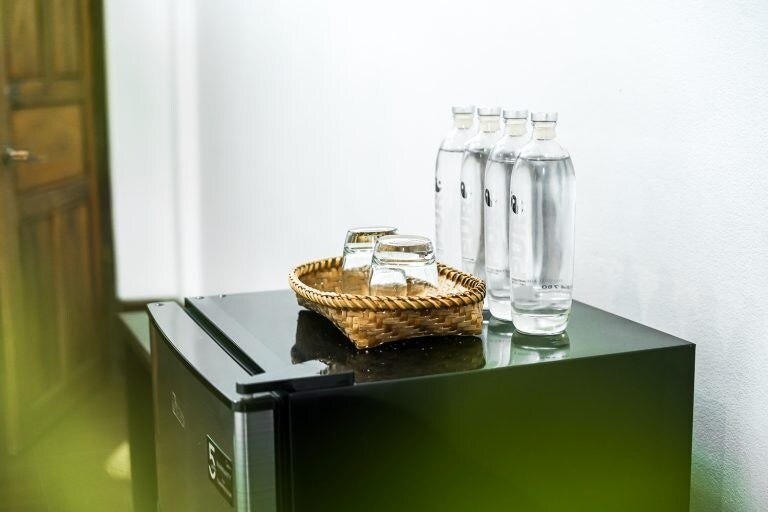 A sleek black refrigerator is positioned against a white wall, topped with a woven tray containing four glass bottles of water and two glasses. Natural light subtly illuminates the scene, enhancing the clean and minimalistic design.
