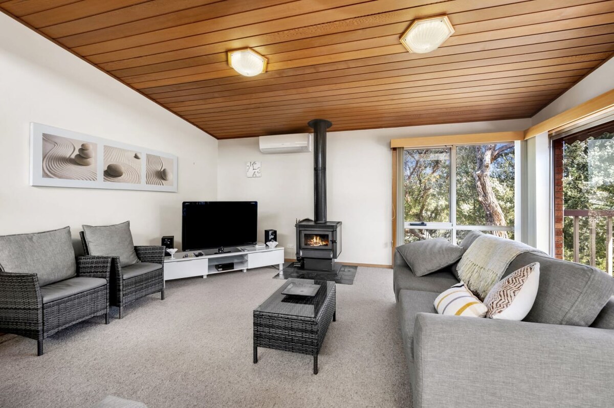 A living area features a cozy arrangement of grey seating, including two armchairs and a spacious sofa. A television is positioned on a white media console, while a wood stove adds warmth. Windows offer views of the surrounding greenery, enhancing the natural light.