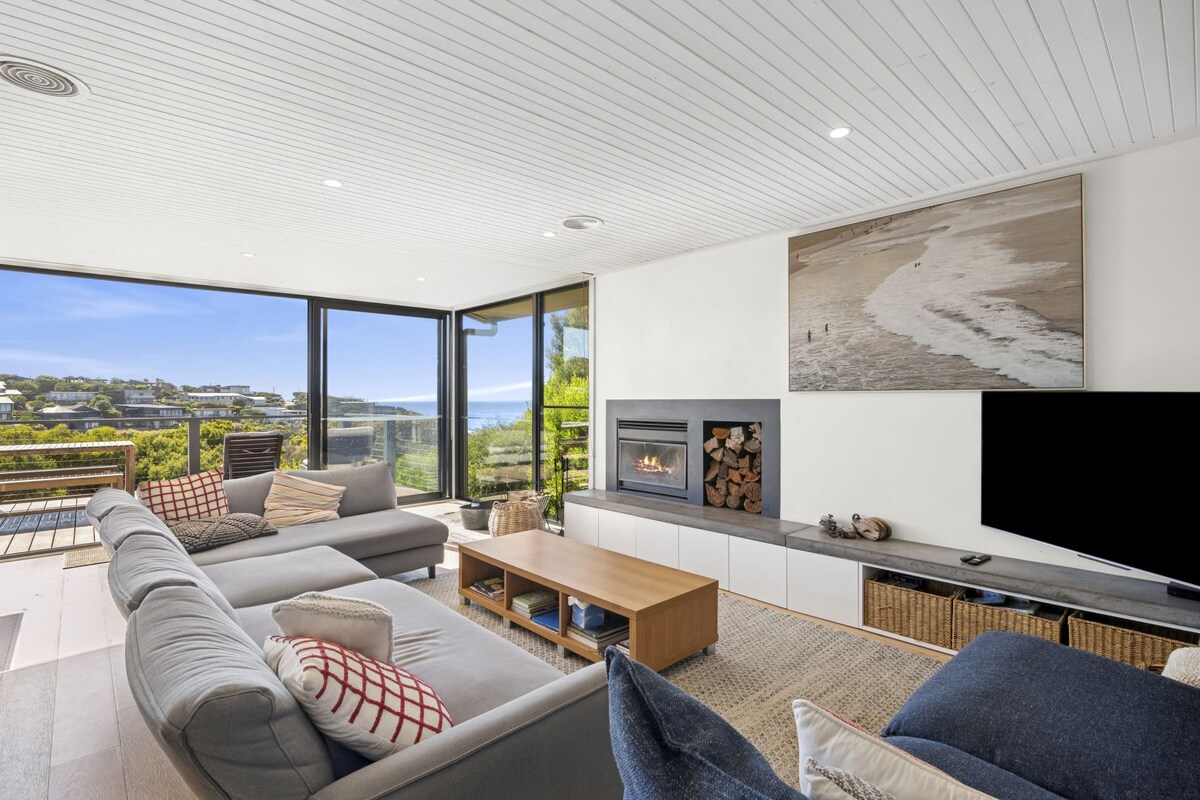 The living area features large glass doors that open to reveal ocean views. A spacious couch is arranged around a wooden coffee table, complemented by a woven area rug. A fireplace is positioned against a light wall, with a piece of art depicting a beach scene.