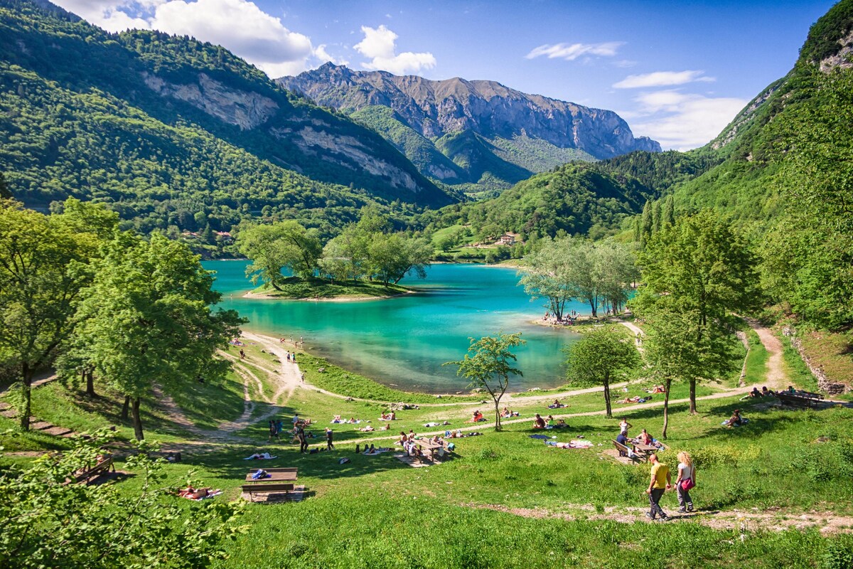 A scenic view showcases the vibrant turquoise waters of Lake Tenno, surrounded by lush greenery and mountainous terrain. Visitors can be seen enjoying the area, with pathways leading through the grassy landscape. Trees provide natural shade along the water's edge, creating a tranquil outdoor setting.