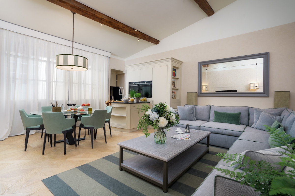 An open-plan sitting and dining area is presented with a spacious L-shaped sofa and a circular dining table accommodating six guests. Natural light filters through sheer curtains, highlighting a neutral color palette complemented by greenery. A stylish kitchen area features integrated appliances, blending comfort with functionality.