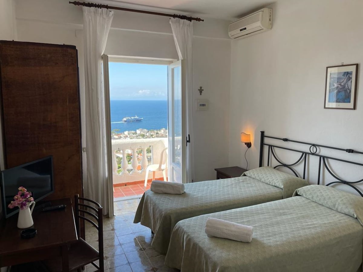 A bright double room is equipped with two spacious beds, a television, and a wooden desk with a chair. Natural light enters through the open sliding door, leading to a panoramic terrace that offers a view of the sea. A hairdryer is positioned on a small shelf.