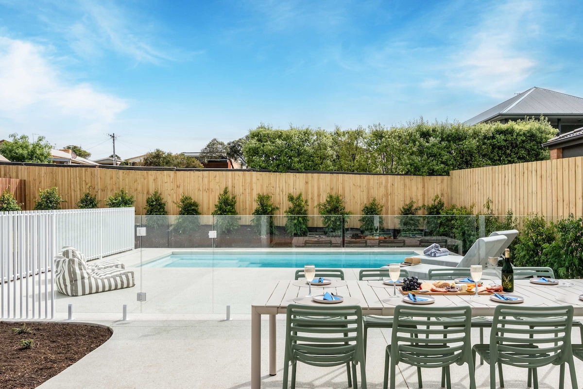 An outdoor dining area is featured next to a swimming pool, complete with a large table and green chairs. A striped lounge chair rests nearby, while lush greenery surrounds the fenced pool area, enhancing the serene setting.