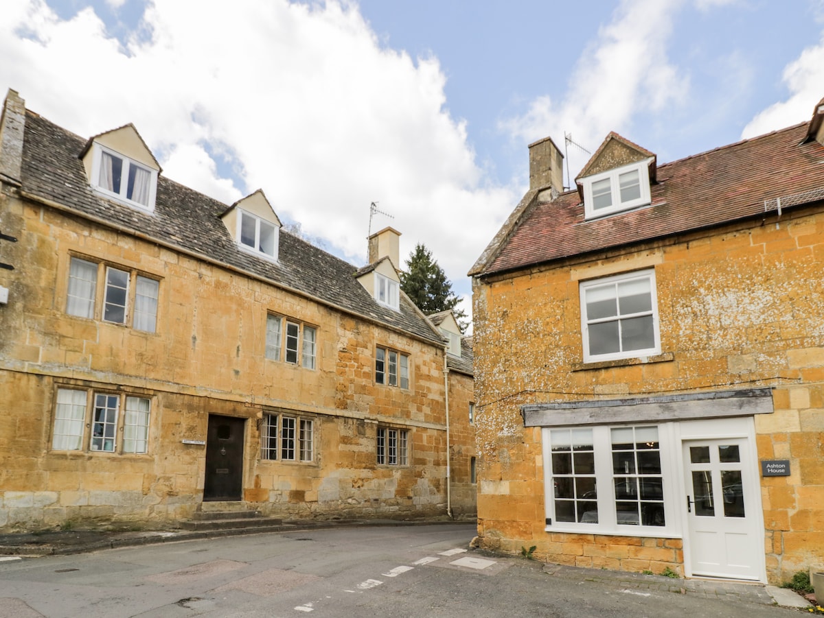 The image displays a charming view of Ashton House, constructed with honey-coloured stone. Its distinctive architecture features multiple gabled roofs and tall windows. The street scene includes a quaint adjacent building, enhancing the village character, with blue skies and scattered clouds above.
