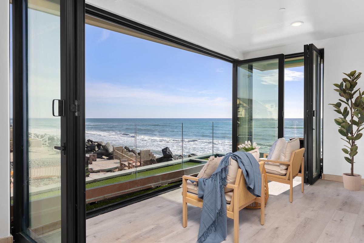 Large glass doors open to reveal stunning ocean views. Natural light fills the open space, featuring two comfortable chairs adorned with a soft blanket. A potted plant adds a touch of greenery, enhancing the fresh coastal ambiance.