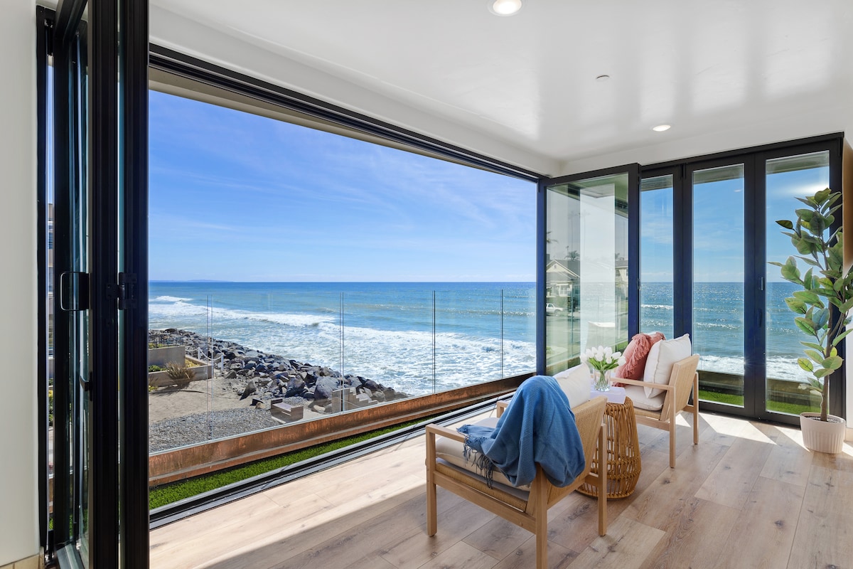 A bright living space features expansive glass walls that open to breathtaking ocean views. Comfortable seating is positioned to face the water, while light wood flooring and greenery enhance the serene atmosphere. Natural light fills the area, creating a relaxed vibe.