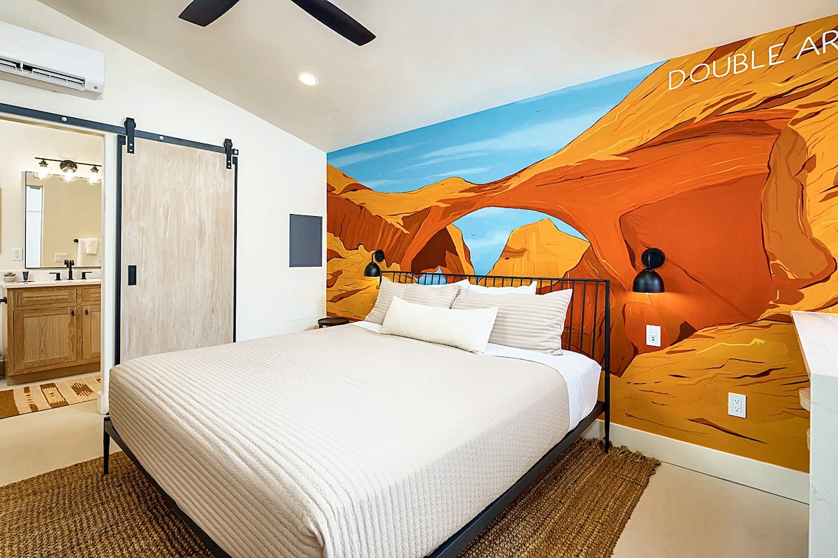 A modern studio features a spacious bed dressed in neutral tones, facing a colorful mural of desert rock formations. Two sleek lamps provide soft lighting on either side. Natural light enters through a window, complementing the elegant decor and adding warmth to the space.