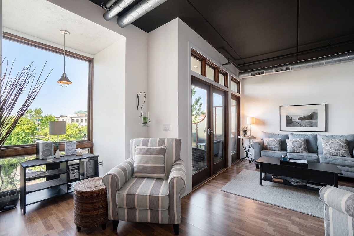 A spacious living area features large windows allowing natural light to brighten the space. A comfortable chair with striped fabric and a woven accent table are positioned beside a modern entertainment unit. The muted color palette and contemporary furnishings create a relaxed environment.