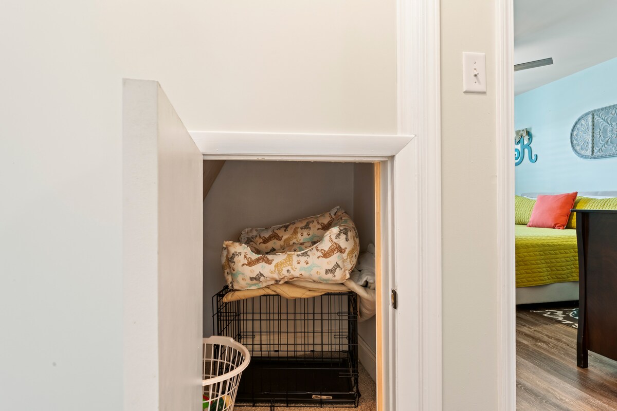 A small storage area is presented with a white door leading to under-stair storage. Inside, a dog crate and a laundry basket are visible, providing practical accommodation for guests' needs. The space is well-organized and easy to access.