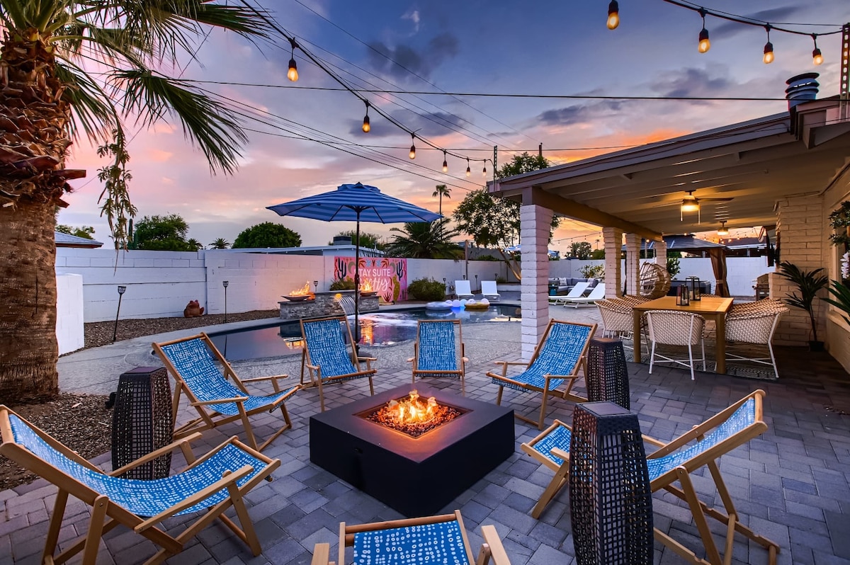 An outdoor space features a cozy fire pit surrounded by eight blue-accented lounge chairs on paved flooring. In the background, a dining area is shaded by an umbrella, with soft lighting strung above. A serene landscape is accented by desert plants and a colorful mural.