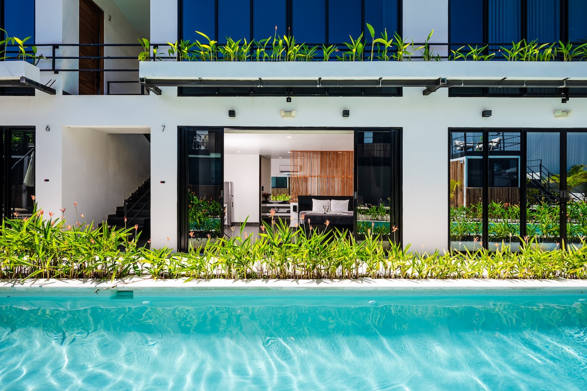 A serene view of the shimmering pool, framed by lush greenery. The minimalist studio with expansive glass doors is visible in the background, seamlessly integrating the indoor and outdoor spaces. The modern design is complemented by natural elements.