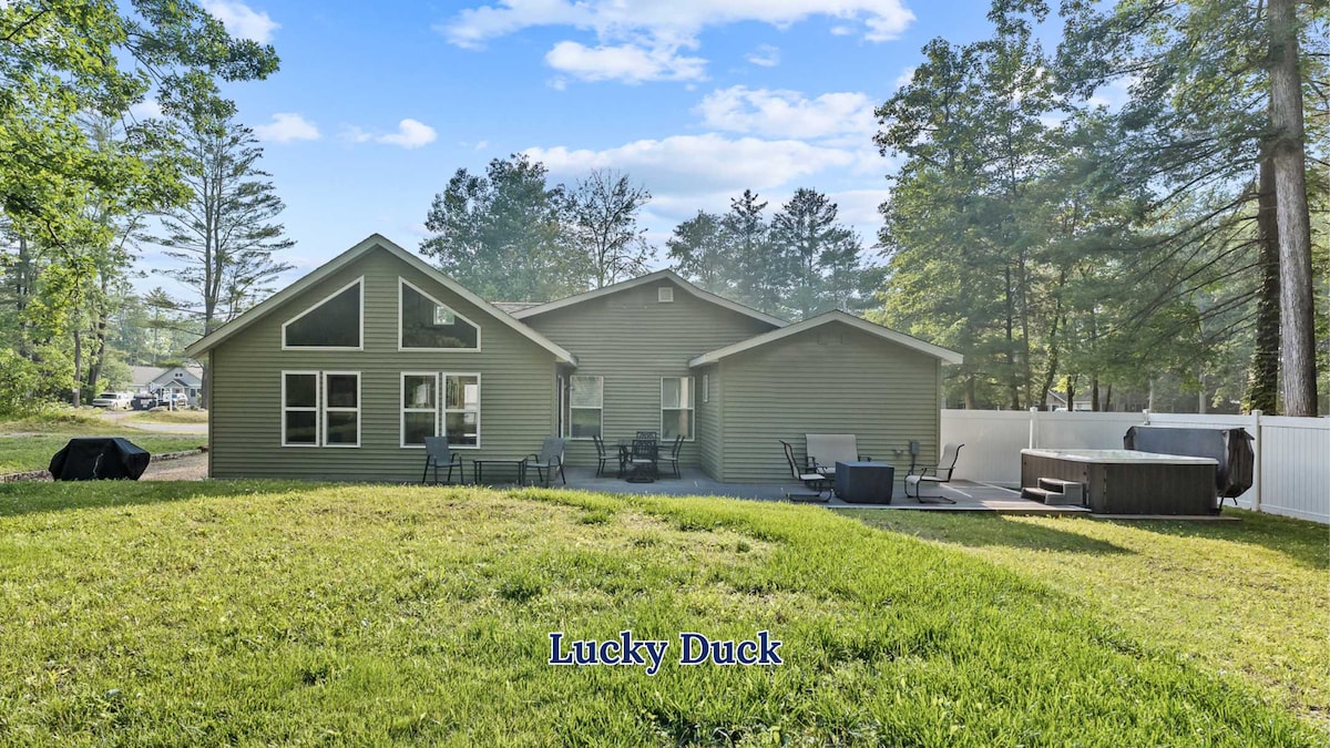 The exterior of Lucky Duck Cottage features a well-maintained green facade surrounded by lush grass. Multiple seating areas are visible, including a patio set and a hot tub. Trees provide a natural backdrop, contributing to the serene ambiance of the backyard.