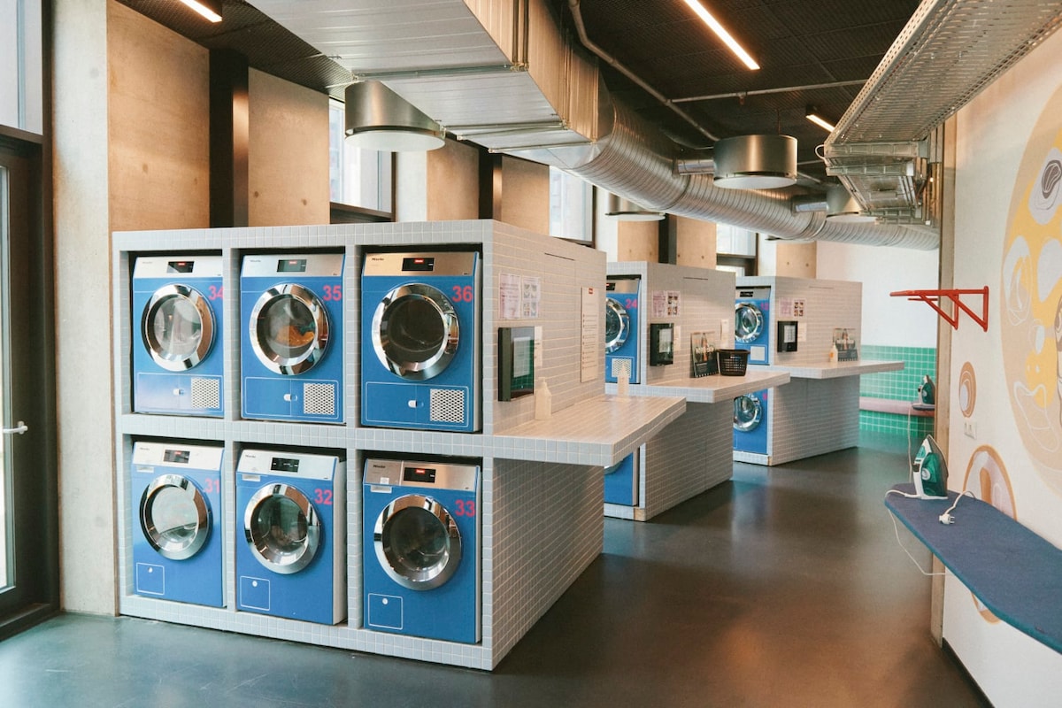 A contemporary laundry facility displays several blue washing machines and dryers lined against the wall. Brightly lit and spacious, the area features countertops for folding clothes and a rolling ironing board, providing a functional and convenient laundry experience.