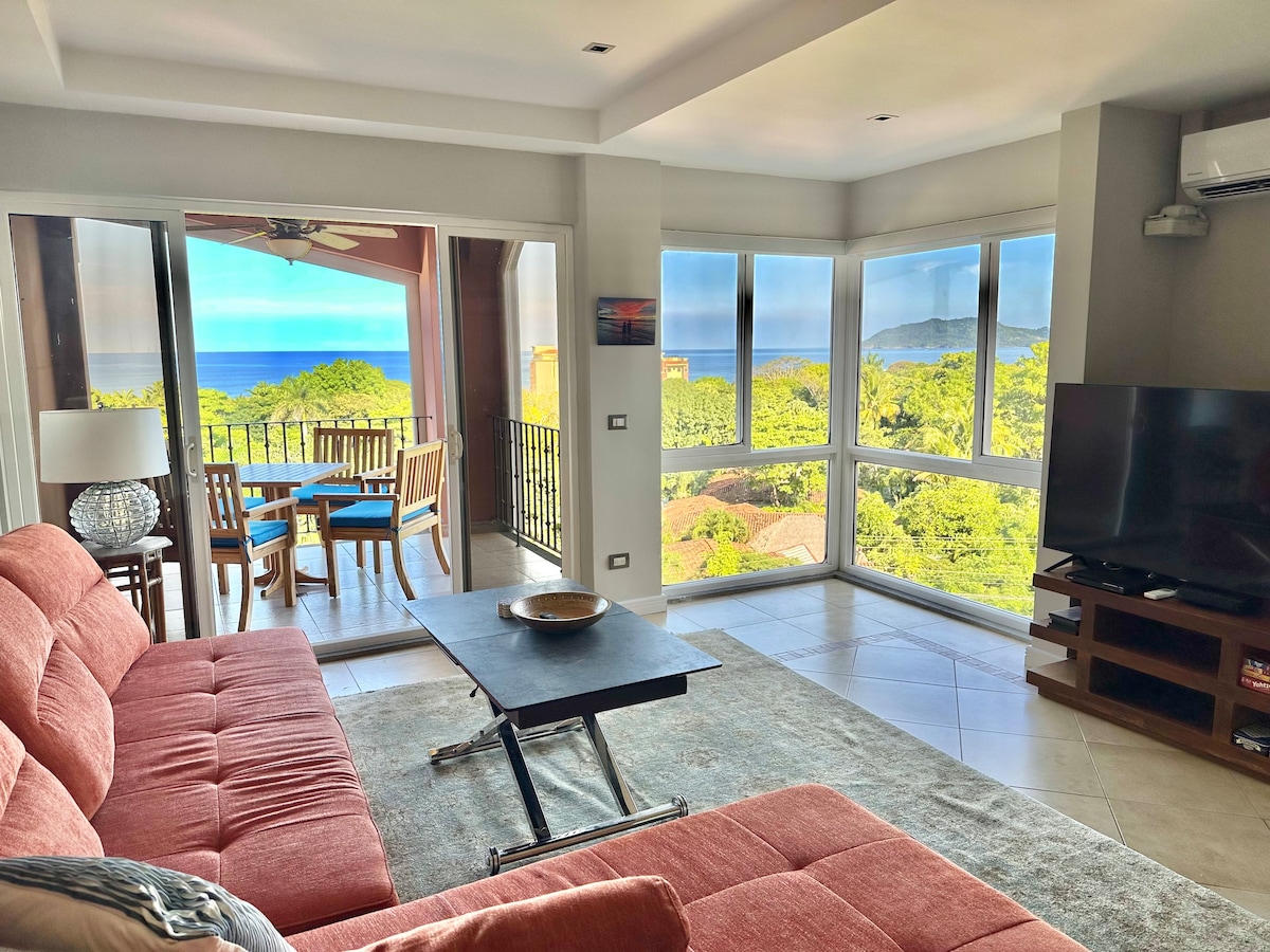 A spacious living area is highlighted by a comfortable orange sofa facing a dark tabletop. Large windows reveal expansive views of the Pacific Ocean and surrounding nature. In the background, a dining area on the terrace includes a wooden table with blue and natural wicker chairs.