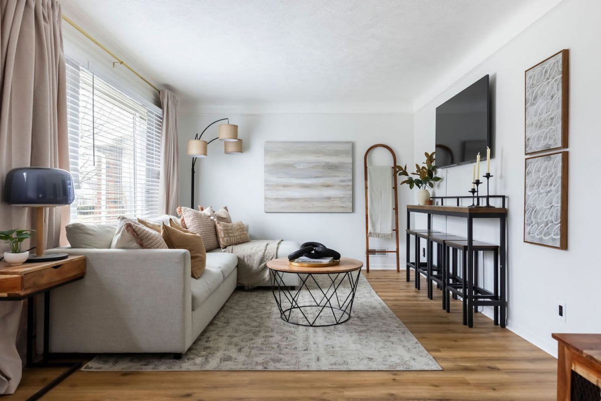 The living room features a spacious sofa adorned with decorative pillows and a large wall-mounted smart TV. Natural light filters through sheer curtains, illuminating the light wood flooring. A circular coffee table sits center stage, complemented by a stylish side table with a lamp.