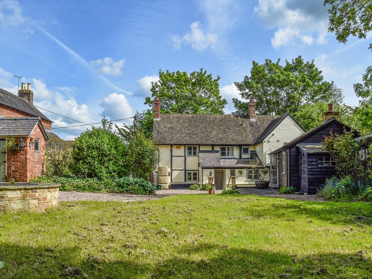 A spacious garden surrounds the Grade II listed cottage, showcasing a well-maintained lawn and a variety of shrubs and trees. The thatched roof cottage, painted in classic colors, stands prominently against a bright blue sky, adding charm to the serene outdoor space.