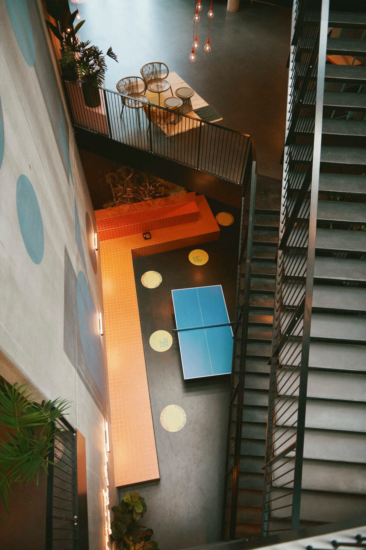 An aerial view showcases a multi-level space featuring a vibrant communal area with seating arrangements and decorative plants. A table tennis setup is visible on a bright orange tiled surface, with circular mats scattered throughout the floor. The staircase is designed with a modern railing, enhancing the open feel.