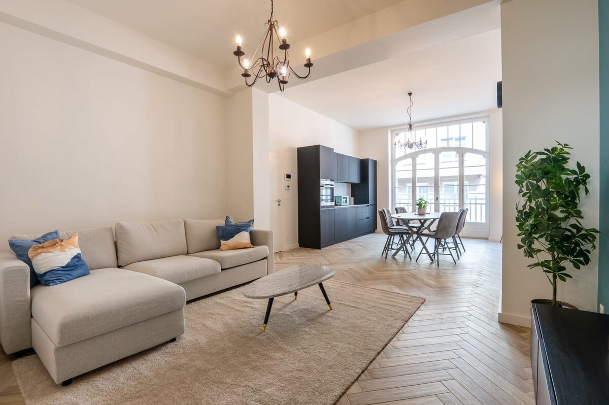 A spacious living area features a light grey sectional sofa adorned with blue and beige cushions. A low coffee table sits on a large area rug, and a dining table with six chairs is visible nearby. The kitchen area showcases sleek dark cabinetry, illuminated by natural light from large windows.