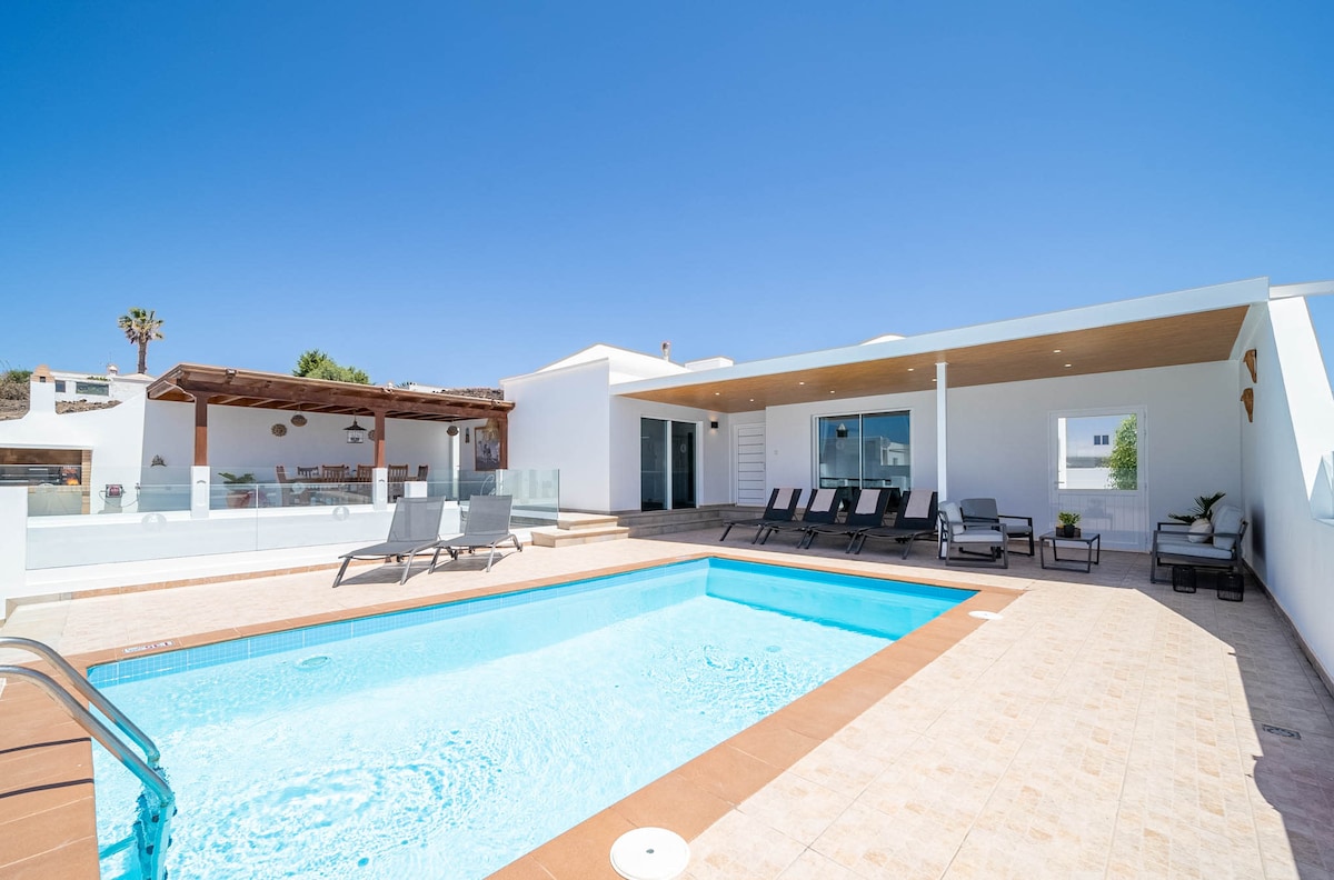An outdoor area features a private pool surrounded by comfortable sun loungers. A spacious terrace with a shaded pergola and dining area overlooks the pool. The modern villa stands in the background, with large glass doors allowing plenty of natural light.