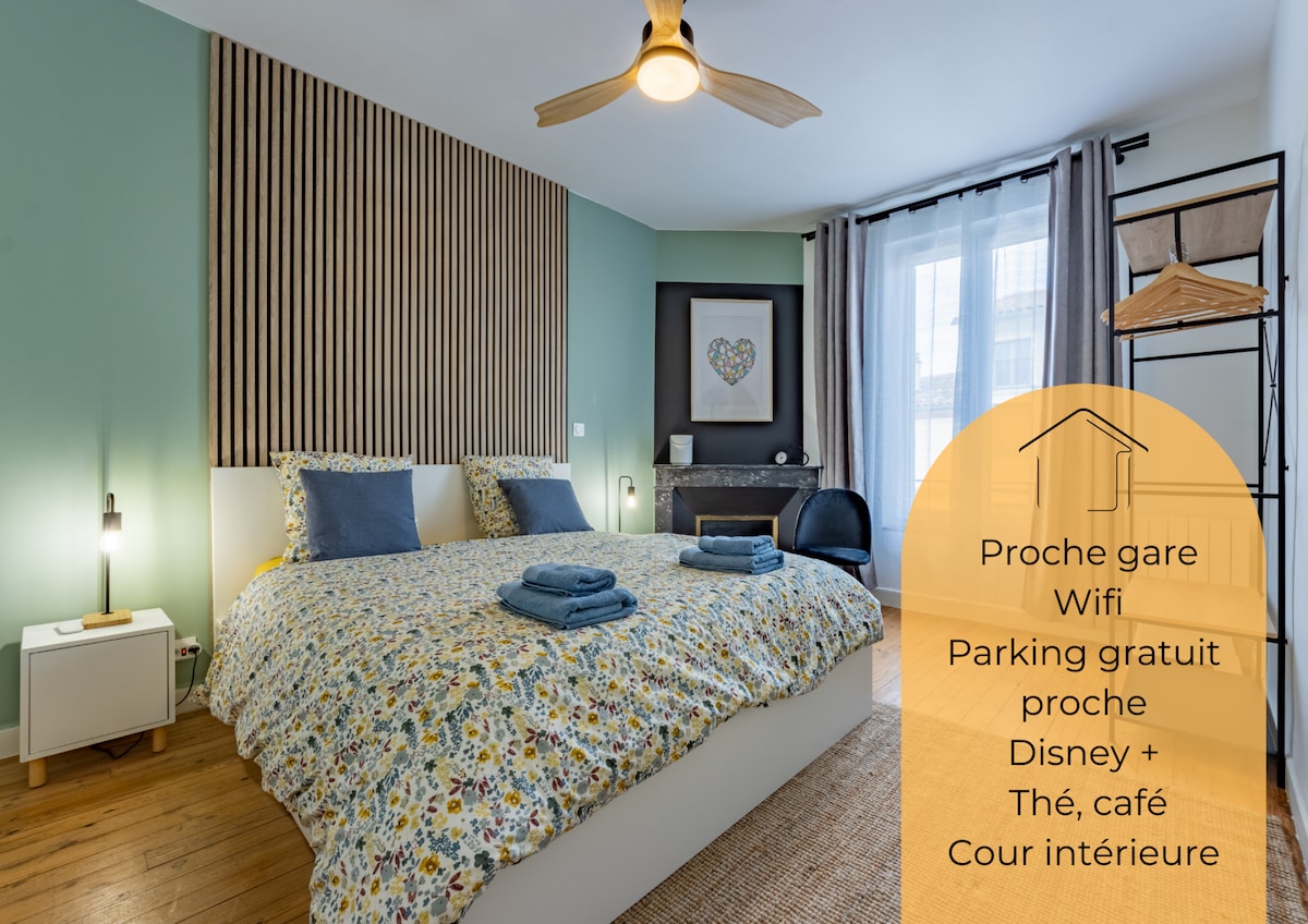 A well-lit bedroom features a spacious bed adorned with a floral comforter. Two towels are neatly placed on the bed. A ceiling fan is present, and natural light enters through the window, complemented by a subtle decorative touch on the wall.