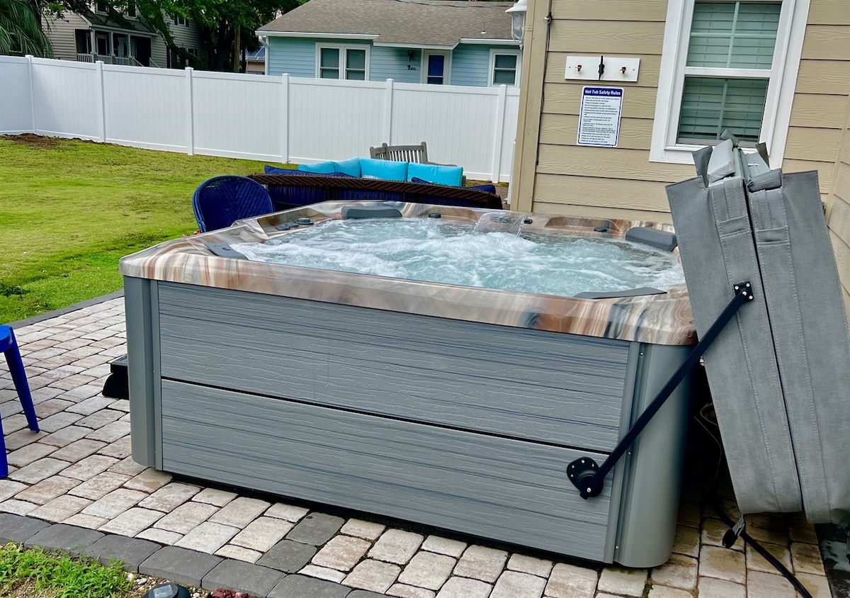 A spacious hot tub is situated on a stone patio surrounded by a manicured lawn. A stylish gray cover is positioned nearby, and blue chairs offer additional seating. The background features a white fence and a glimpse of the home.
