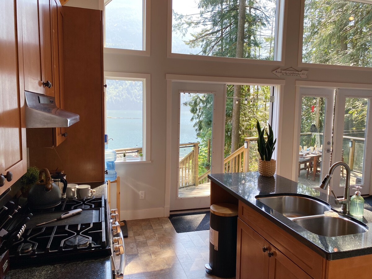 The kitchen features modern wooden cabinetry with a dark countertop and a gas stove. Large windows offer a view of the lake and trees, allowing natural light to fill the space. A sliding glass door opens to a deck, enhancing the connection to the outdoors.