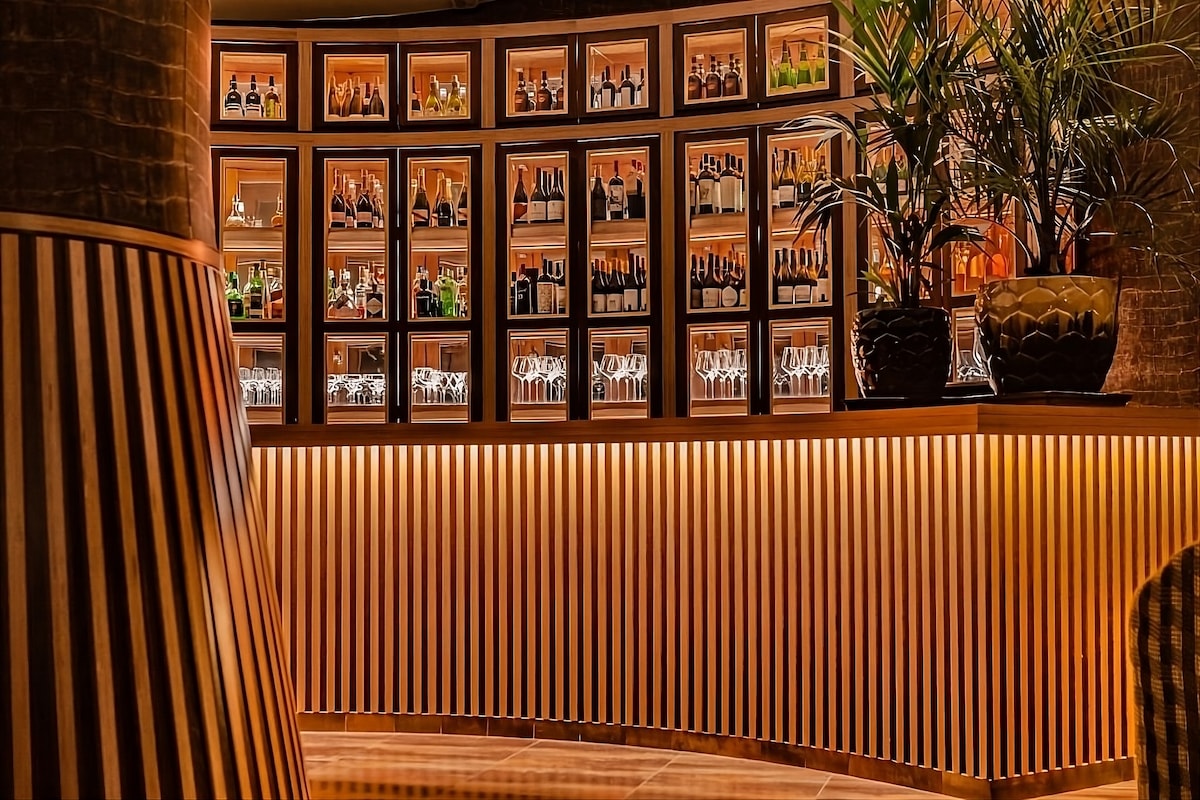 A well-organized bar area features a curved wooden counter illuminated by warm lighting. Shelves behind the counter are lined with various bottles and glassware, complemented by decorative plants in elegant black pots.