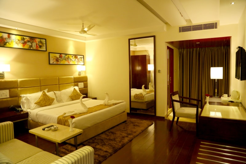 A well-furnished room features a spacious bed with decorative pillows. A mirrored closet reflects the soft lighting, while a cozy sitting area includes a wooden chair and a small table. Decorative artwork is displayed above the bed, and a window with curtain offers privacy.