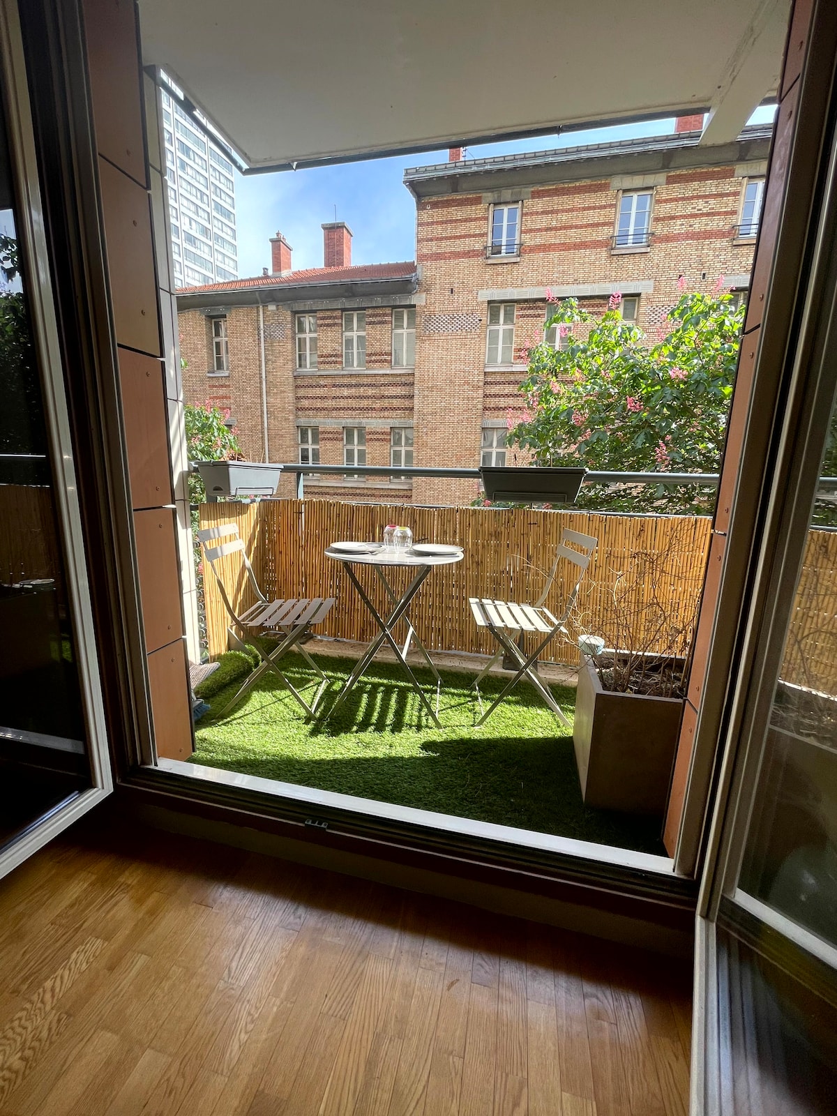 The image captures a sunny balcony space featuring a small round table and two chairs, surrounded by green plants. The backdrop consists of a brick building, with sunlight illuminating the area, creating a welcoming atmosphere for outdoor relaxation.