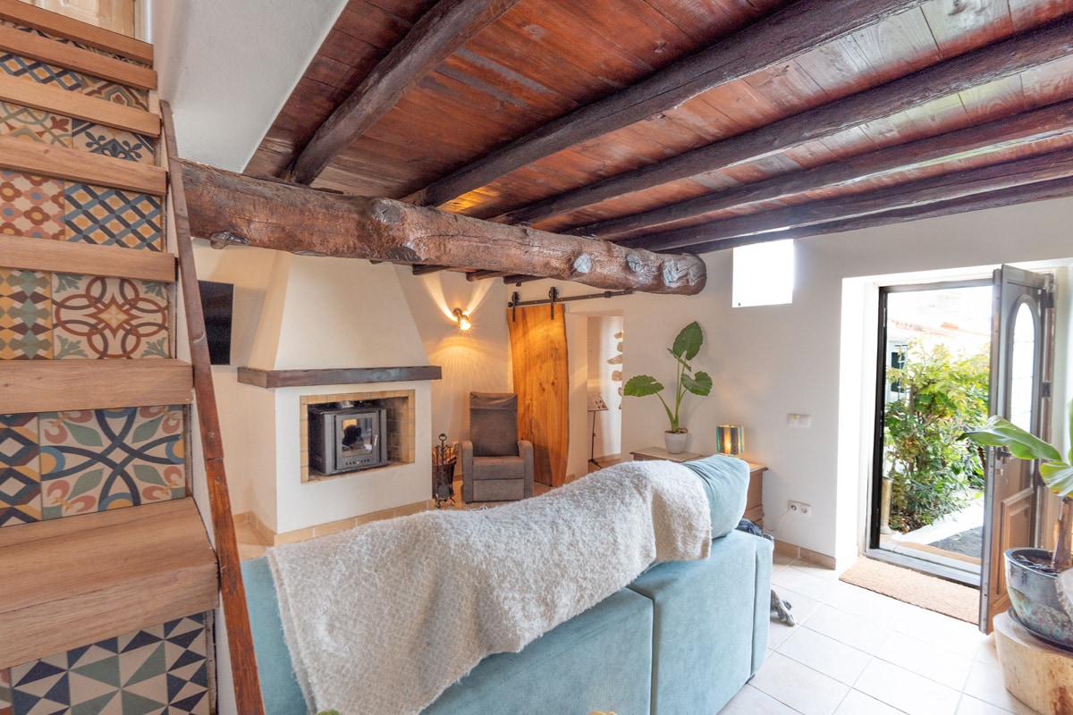A cozy living area features wooden beams and a soft-color sofa draped with a light blanket. A modern fireplace is positioned against one wall, complemented by a small armchair. Indoor plants add a touch of greenery, while natural light filters in from a window.