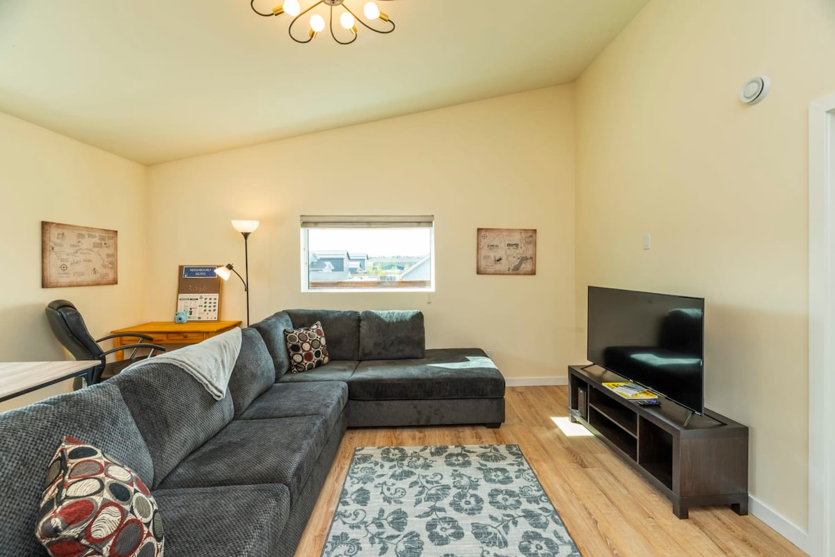 A spacious living area is furnished with a large sectional couch adorned with patterned pillows. An entertainment unit houses a television, and a small desk with a lamp is visible in the corner. Natural light filters in through a nearby window, enhancing the cozy atmosphere.