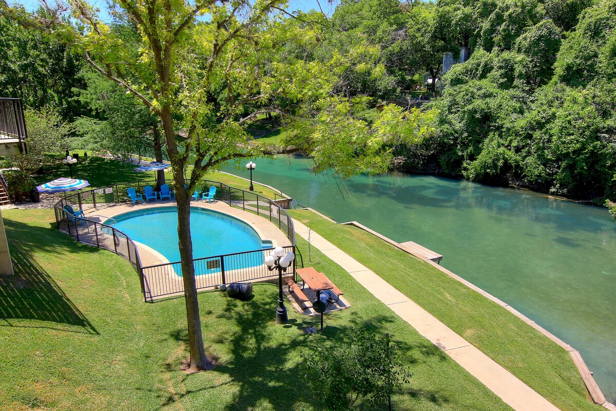 An outdoor area is visible featuring a swimming pool surrounded by a fence. Lounge chairs are arranged nearby, and picnic tables are positioned on the grass. Direct river access is shown, along with lush greenery along the banks.