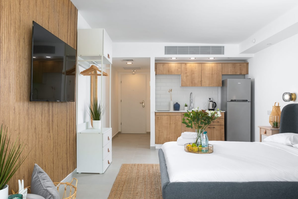 A studio apartment features light wooden cabinetry and an equipped kitchenette. A spacious bed with crisp white linens is positioned next to a television mounted on the wall. Decorative plants add a touch of greenery, enhancing the overall welcoming ambiance of the space.