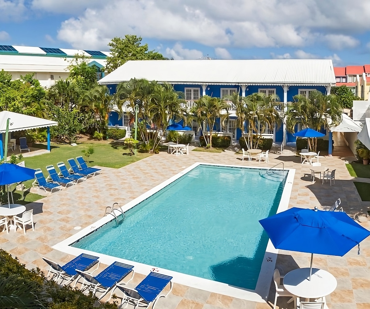 A large swimming pool is centered in a landscaped outdoor area, surrounded by sun loungers and umbrellas. The pool area is accented by tropical greenery and features a spacious deck with several seating options, promoting a relaxing atmosphere.