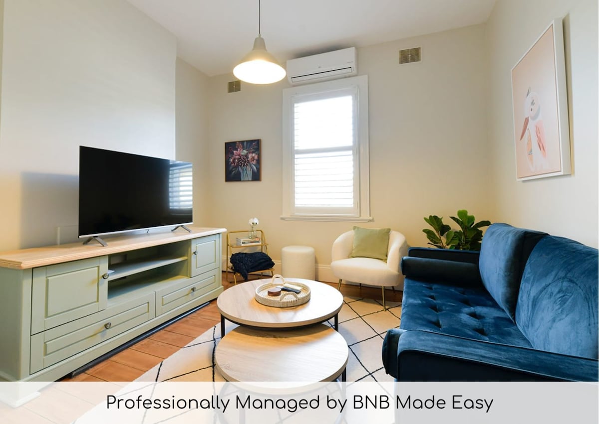 A welcoming living area features a blue sofa and an accent chair, providing a comfortable spot to relax. A television is positioned on a light wooden console, while a round coffee table sits at the center. Natural light enters through the window, enhancing the serene ambiance.