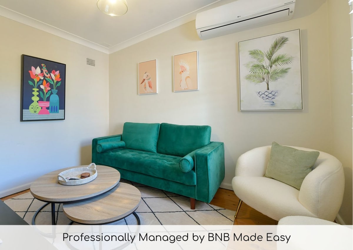 A bright living area features a green sofa, complemented by a light-colored armchair. Framed artwork adorns the walls, adding color to the space. A round coffee table stands in the center, enhancing the cozy atmosphere.