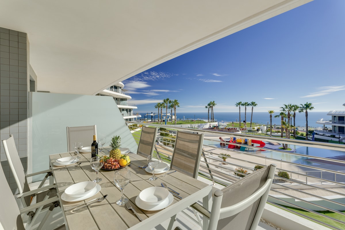 A spacious terrace overlooking the complex features a dining table set for six, complete with plates and wine glasses. The view includes palm trees, swimming pools, and glimpses of the sea in the background, creating a relaxed outdoor dining area.