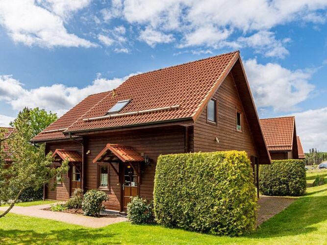 A two-story, wooden holiday home is surrounded by neatly trimmed hedges and lush greenery. The sloped roof features a skylight, and the entry is adorned with two small awnings. Bright clouds drift across a blue sky, highlighting the inviting exterior of the property.