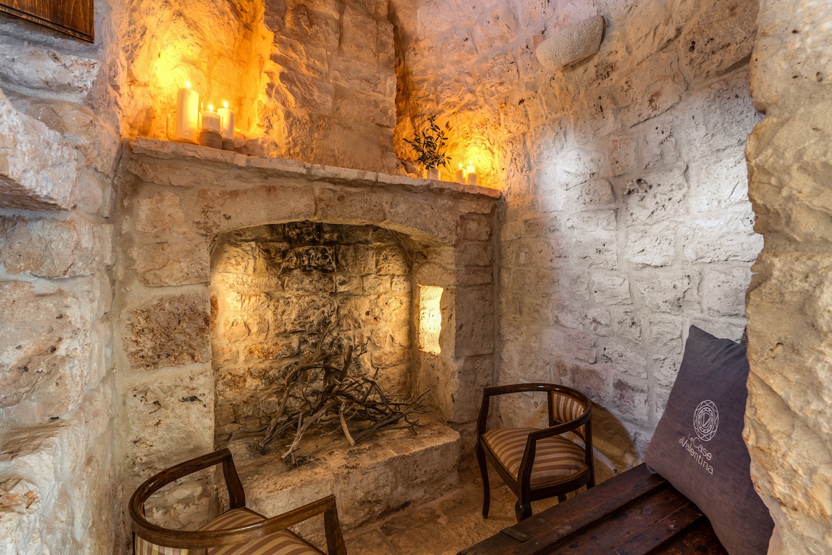 A cozy seating area is highlighted by a rustic stone fireplace, its warm glow accentuated by candles placed on the surrounding ledge. Two wooden chairs are positioned near the fireplace, creating an inviting atmosphere for relaxation.