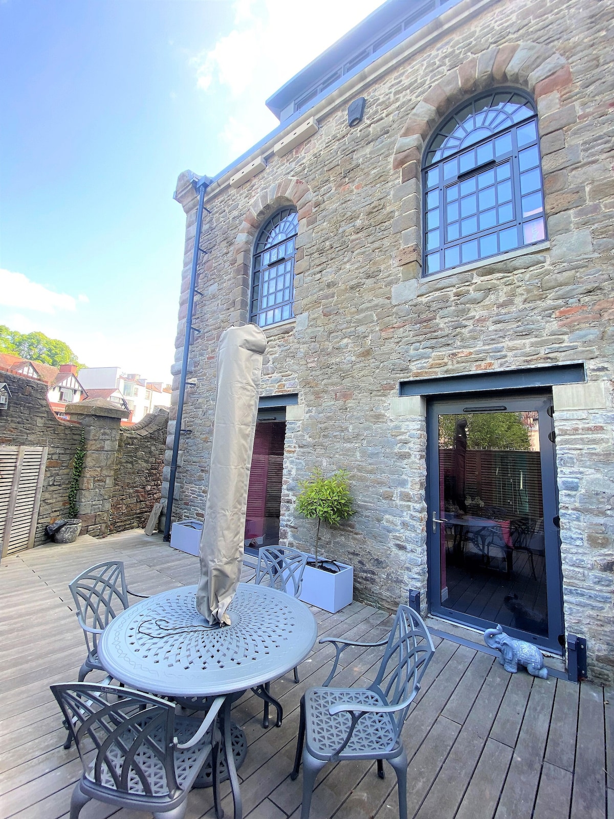 The outdoor terrace features a spacious wooden deck with a round table surrounded by six matching chairs. Two tall windows with arched tops are visible on the stone wall, complementing the modern glass doors that lead inside.