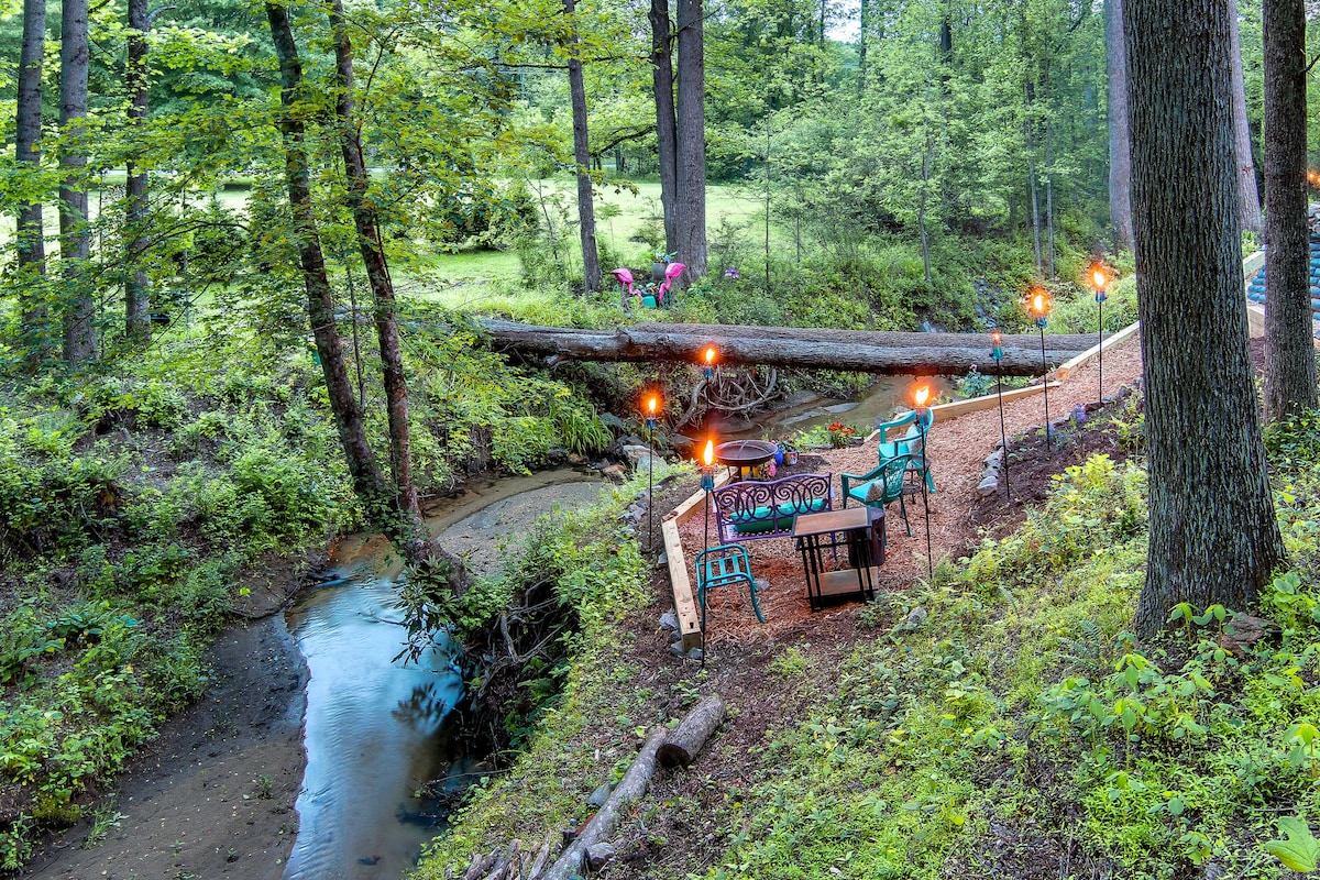 An outdoor seating area is positioned beside a winding creek, featuring a table and chairs with amber lights strung above. Lush greenery surrounds the space, and a fallen log crosses the creek, enhancing the natural setting.